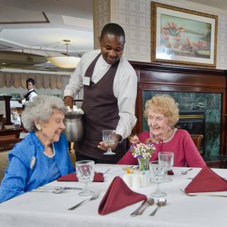 A waiter serves two elderly women seated at a dining table in a restaurant.