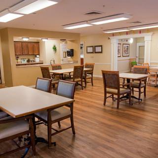A well-lit dining area with wooden flooring, furnished with tables and chairs, adjacent to a kitchenette.