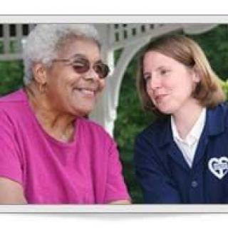 An elderly woman and a caregiver share a smile outdoors.