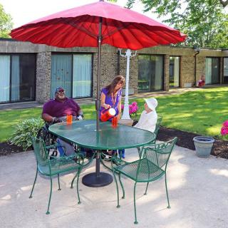 Three people sitting at a patio table under a red umbrella in a garden setting.