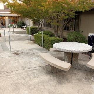 A round outdoor table with attached benches situated on a paved area near trees and a pathway.