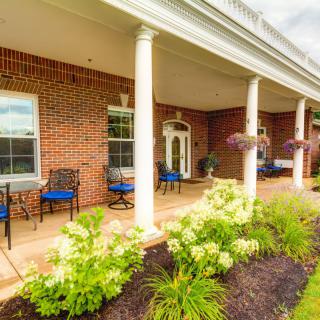 A charming brick building with a porch featuring white columns, neatly arranged chairs, and vibrant landscaping.