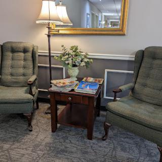 A cozy living room setting featuring two green armchairs, a wooden side table with magazines, and a lamp, complemented by a vase of flowers.