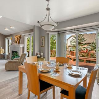 A modern dining area with a wooden table set, adjacent to a cozy living room illuminated by abundant natural light from large windows.