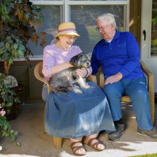 An elderly couple sitting on a porch, smiling as they enjoy time with their dog.