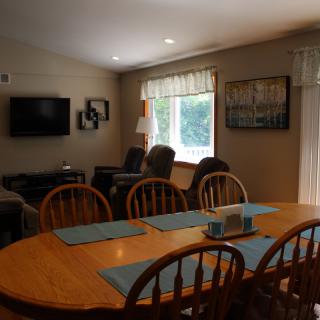 A cozy dining and living area featuring a wooden dining table with chairs, a mounted television, armchairs, and a window with curtains.