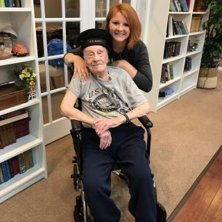 A woman smiling and posing with an elderly person seated in a wheelchair, set in a cozy library environment.
