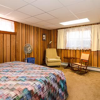 A cozy basement bedroom with wood paneling and a quilt-covered bed, featuring an armchair, rocking chair, and standing fan.