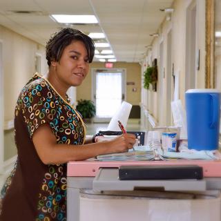 A nurse standing in a hospital corridor holding a clipboard.