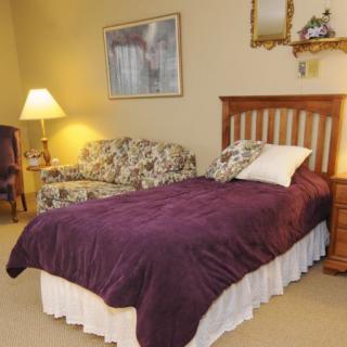 A cozy bedroom with a single bed covered in a purple blanket, accompanied by a floral sofa and a lamp.