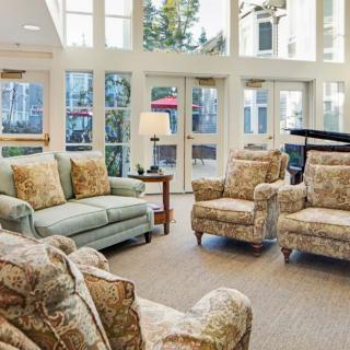 A cozy living room with plush sofas and chairs, a piano, large windows, and a lamp adding warmth to the space.