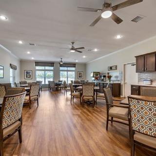 A spacious dining area with wooden floors, multiple tables and chairs, ceiling fans, and large windows.