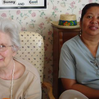 An elderly woman sitting with a caregiver, both smiling, in a cozy indoor setting.