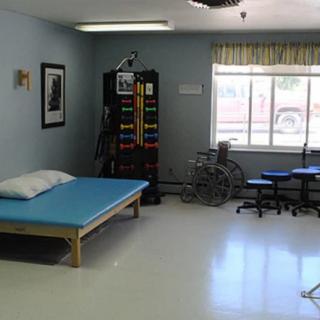 A physical therapy room equipped with a treatment table, exercise equipment, and a wheelchair by the window.