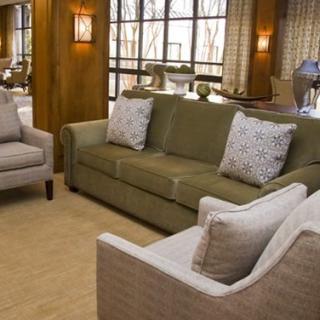 A cozy living room with a green sofa and two beige armchairs, complemented by patterned cushions and warm wood paneling.