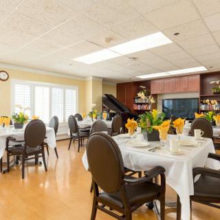 A well-lit dining area with neatly arranged tables and chairs, decorated with yellow flowers. A piano is visible in the background.