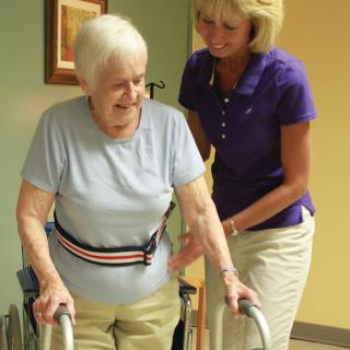 A caregiver assists an elderly woman using a walker during a physical therapy session.