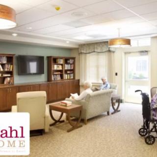 A cozy living room area in a nursing home with comfortable seating, a television, and bookshelves.