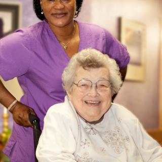 A caregiver standing behind an elderly woman sitting in a wheelchair, both smiling warmly.