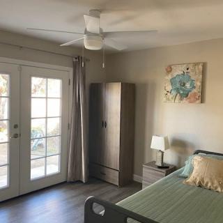 A cozy bedroom with a modern interior, featuring a comfortable bed, wardrobe, bedside lamp, ceiling fan, and double doors leading outside.