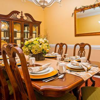 A warmly lit dining room with a wooden table set for a meal, adorned with elegant tableware and a flower arrangement.