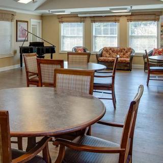 A community room with tables, chairs, and a piano near large windows.