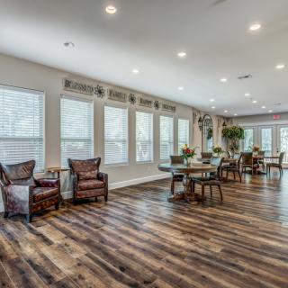 A spacious living room with wooden flooring and ample natural light shining through large windows, furnished with chairs and tables.