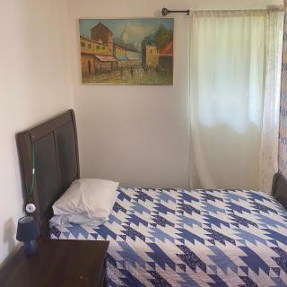 A cozy bedroom with a single bed featuring a blue and white patterned bedspread, a pillow, a bedside table, a painting on the wall, and a window with white curtains.