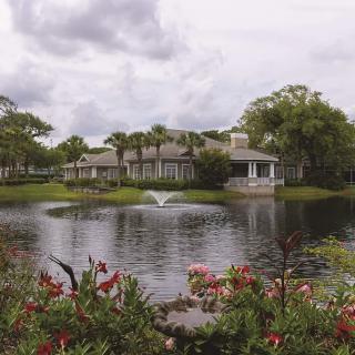 A large house by a tranquil lake with a fountain, surrounded by lush gardens and trees under an overcast sky.