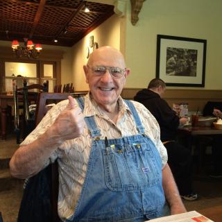 A cheerful man in overalls gives a thumbs-up at a restaurant.