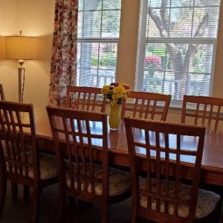 A dining room with a large wooden table and chairs, a vase of flowers, and natural light coming through the windows.