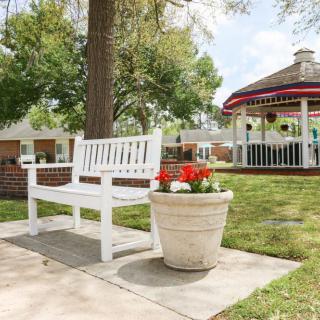 A white bench and a potted plant with red flowers sit on a paved walkway near a gazebo in a lush garden setting.