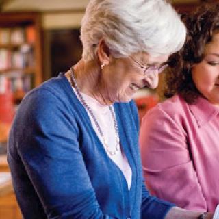 An elderly woman and a younger woman engage in a warm interaction while exploring technology together.