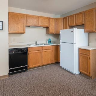 A modern apartment kitchen with wooden cabinets, a refrigerator, and carpeted flooring.