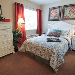 A cozy bedroom with a neatly made bed, decorated with floral artwork and bright curtains.