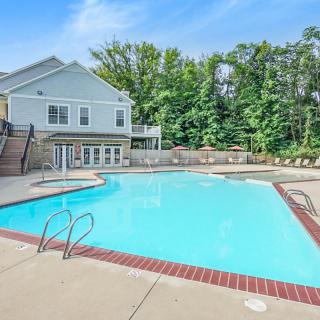 A serene outdoor swimming pool adjacent to a residential house, surrounded by lush trees and a clear blue sky.