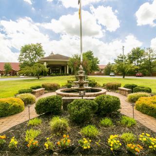 A beautifully landscaped garden with a central fountain surrounded by lush greenery and colorful flowers under a cloudy sky.