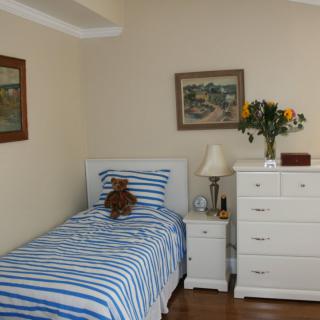 A cozy bedroom with a single bed featuring striped bedding, and a dresser topped with flowers and a lamp.