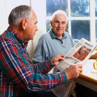 Two elderly men sitting at a table, one reading a newspaper.