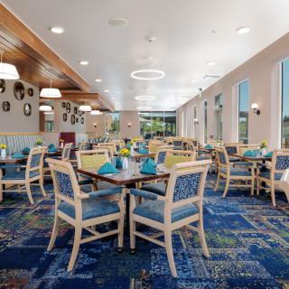 A modern dining room with neatly arranged tables and chairs, featuring a patterned carpet and ample natural light.
