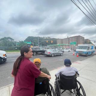 A caregiver assisting two individuals in wheelchairs on a busy city street under a cloudy sky.