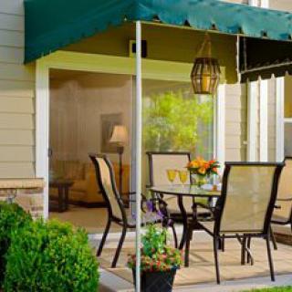 A cozy patio area with a table and chairs under a green awning, surrounded by lush greenery and flowers.
