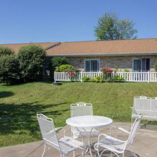 A sunny outdoor patio with a white table and chairs, overlooking a well-maintained garden and lawn in front of a house.