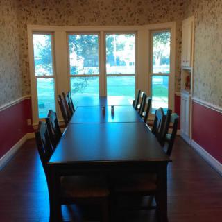 A dining room featuring a long table with chairs, set against large windows.