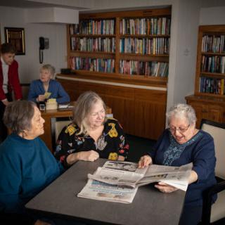 A group of seniors gathered around a table, enjoying a lively conversation while reading newspapers in a cozy library setting.