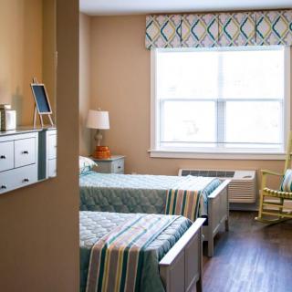 A well-lit hospital room with twin beds, featuring a lamp on the nightstand and a rocking chair by the window.