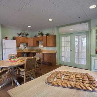 A cozy kitchen with a dining area, featuring a table set with cookies, surrounded by wooden cabinets and indoor plants.
