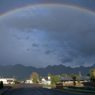 A stunning rainbow arches over a quiet road, with mountains in the background and dark clouds hinting at recent rain.