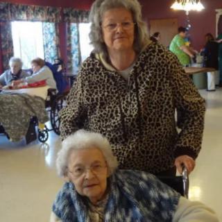 A middle-aged woman stands behind an elderly woman in a wheelchair inside a nursing home setting.