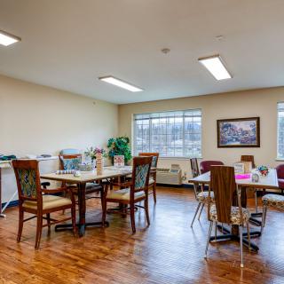 A bright dining room with several tables and chairs, featuring large windows and artwork on the walls.